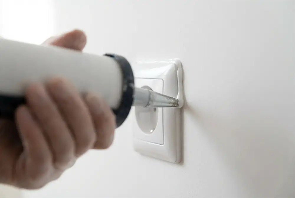 Gros plan sur l'application de mastic acoustique autour d'un boîtier électrique pour stopper les fuites d'air et le passage du son.
