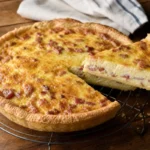 Quiche lorraine authentique sans fromage, bien dorée, posée sur une grille, avec une tranche découpée révélant la migaine crémeuse aux lardons